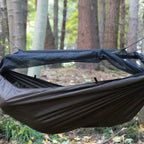 DD Frontline Hammock i Coyote Brown hengt opp i en skog, tett på, med myggnettet åpnet på den ene siden og lagt opp over spilen