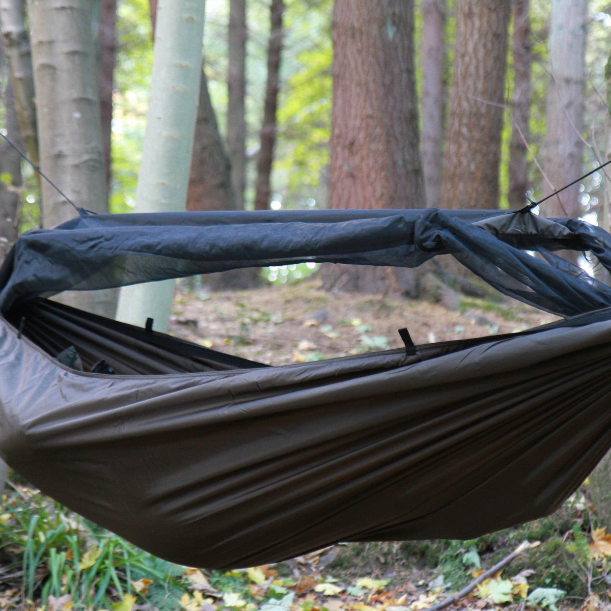 DD Frontline Hammock i Coyote Brown hengt opp i en skog, tett på, med myggnettet åpnet på den ene siden og lagt opp over spilen