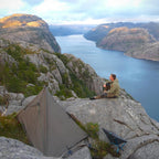 DD Tarp 3x3 i Coyote Brown satt opp som telt på fjellet over en fjord, med en mann som sitter på en stein ved siden av
