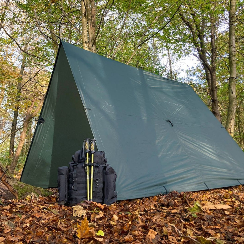 DD Tarp XL i Olivengrønn satt opp som telt ned til bakken, med en sekk og to staver foran åpningen